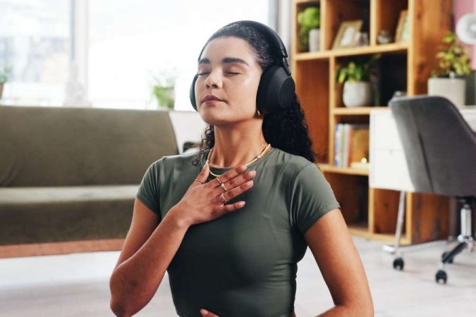 Femme pratiquant des techniques de respiration à domicile, main sur la poitrine, yeux fermés et casque audio, dans un salon lumineux et apaisé.