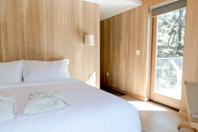 Une chambre lumineuse aux murs et plafond en bois clair avec un grand lit défait, un peignoir posé dessus et une porte vitrée donnant sur une forêt.