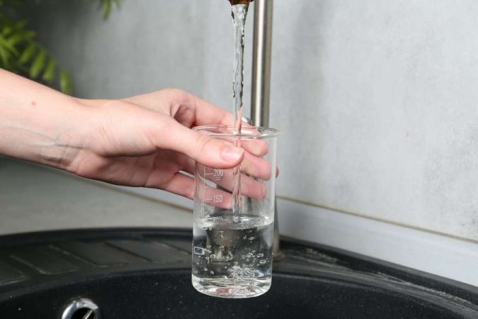 Une main tient un verre gradué sous un robinet de cuisine pour mesurer le débit et la pression d'eau.